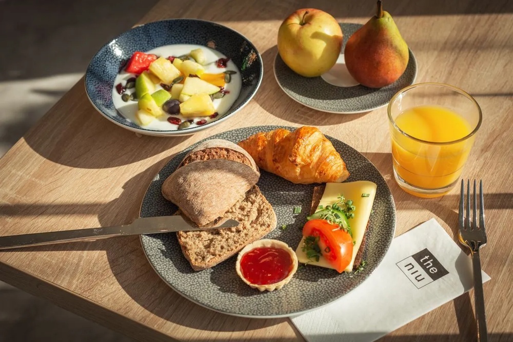 Ein Frühstücksteller mit Croissant, Brot, Käse, Tomate, Marmelade, Müsli, Apfel, Birne und Orangensaft.
