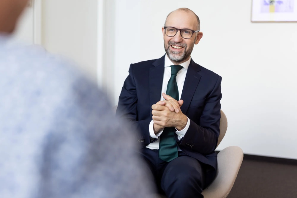 Ein lächelnder Mann in Anzug und Krawatte sitzt auf einem Stuhl in einem Büro.