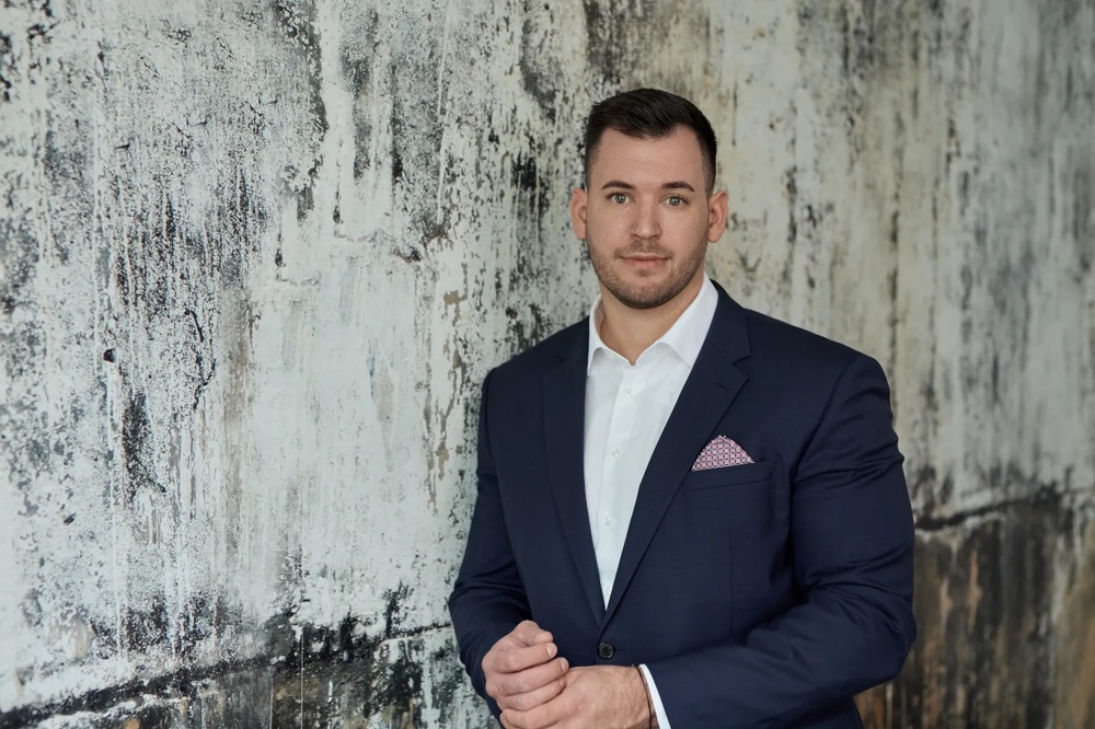 Ein Mann trägt einen schwarzen Anzug ohne Krawatte und ein Einstecktuch. Er steht vor einer marmorierten Wand.