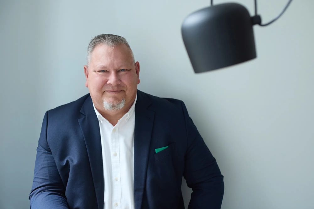 Es wird der Commerz Real Mitarbeiter Claus André Krüger, Senior Key Account Manager für Sales Private and Business Clients SW, dargestellt. Er sitzt vor einer hellgrauen Wand mit einer schwarzen Deckenleuchte, trägt ein dunkelblaues Sakko und blickt ruhig in die Kamera.