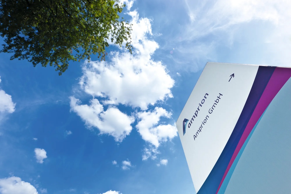 Ein Schild mit "Amprion GmbH" vor blauem Himmel mit weißen Wolken und einem Baum.