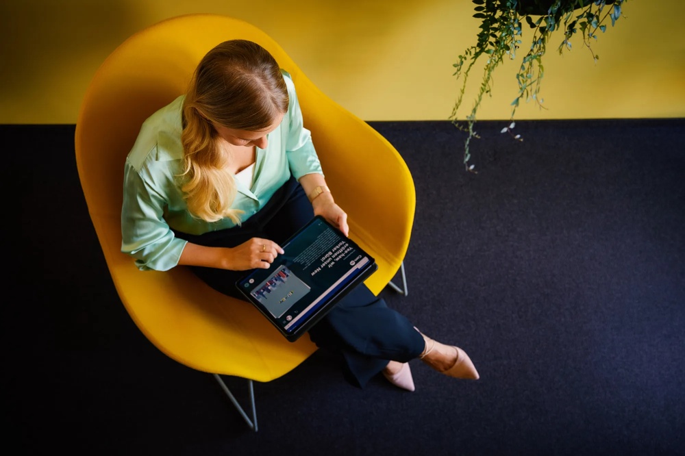 Woman in yellow chair looking at tablet, green shirt, yellow background, hanging plant.