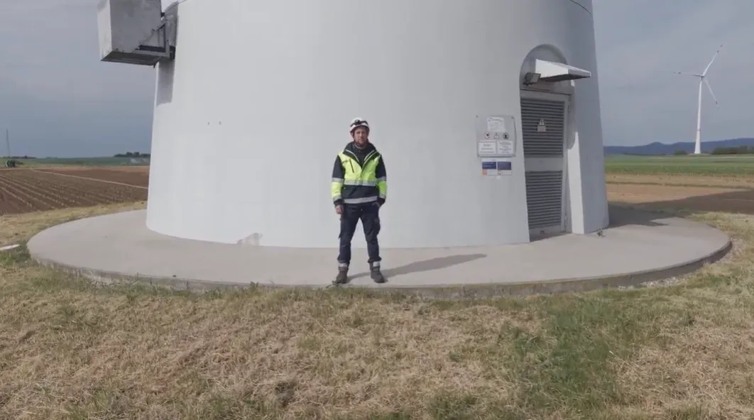 Wartungstechniker mit Helm und neonfarbener Jacke vor dem Sockel eines Windkraftrads.