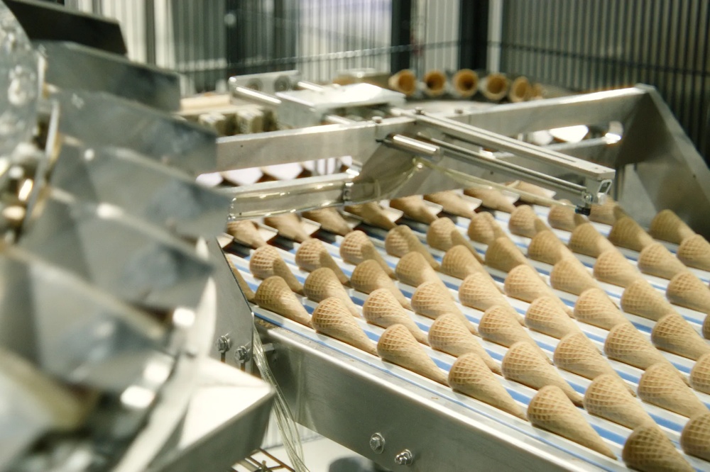 Zwei Fabrikationsmitarbeiter stehen in weißen Kitteln gebeugt über einer Eiswaffelproduktionsstraße und begutachten die Reihen der frischgebackenen Hörnchen.
