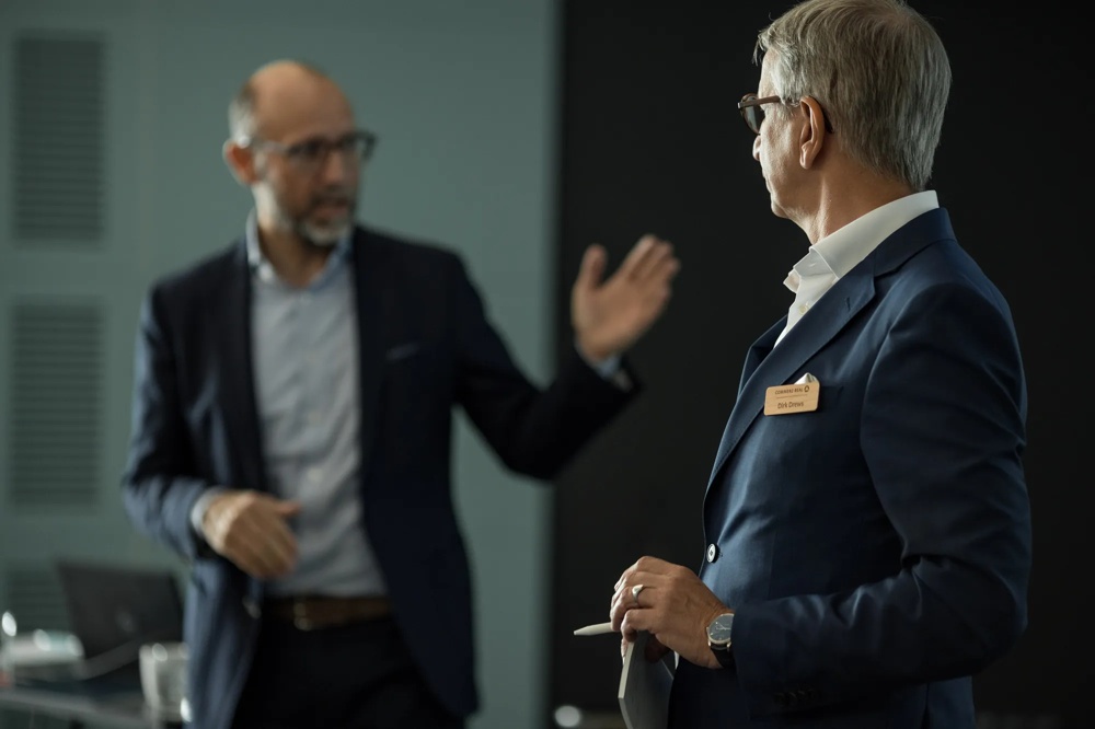 Zwei Männer in Anzügen diskutieren in einem Büro, einer hält Papiere, der andere gestikuliert.