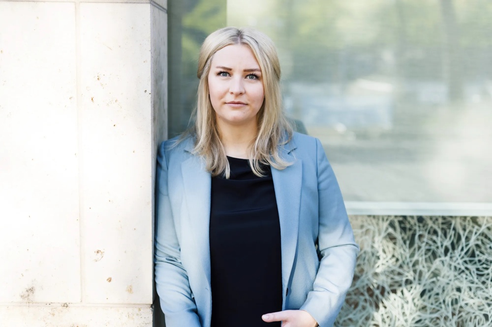 A woman in a blue blazer standing in front of a window looking into the camera.