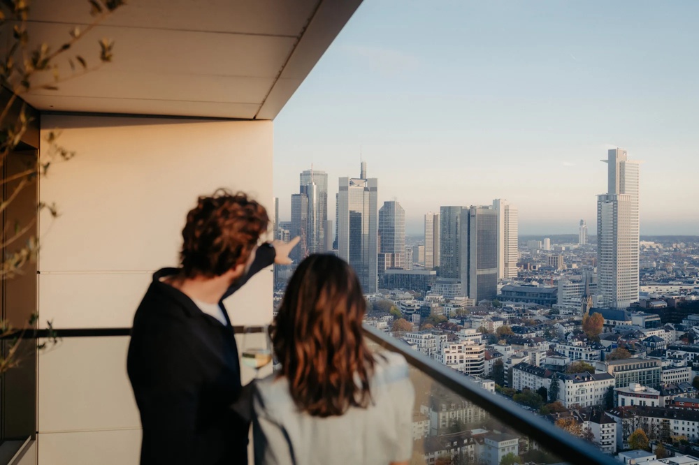 Zwei Personen auf Balkon einer modernen Wohnung mit Blick über die Stadt.