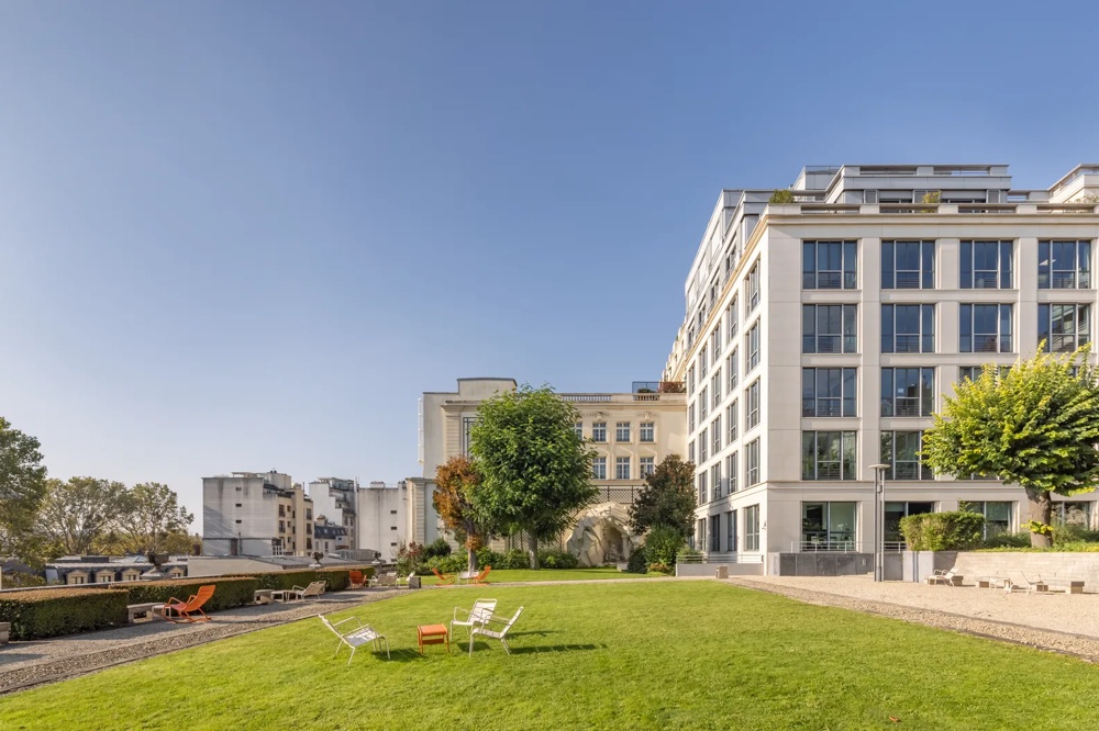 Grüne Wiese mit weißen Stühlen, modernem Gebäude und Eiffelturm im Hintergrund bei klarem Himmel.