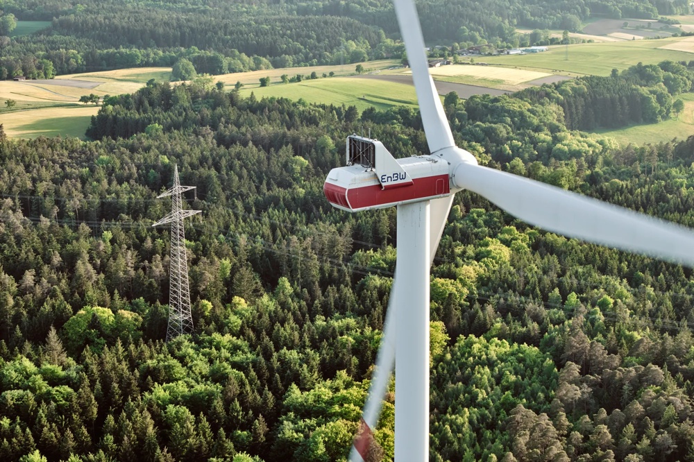 Drei Windräder stehen in einem grünen Wald unter blauem Himmel.