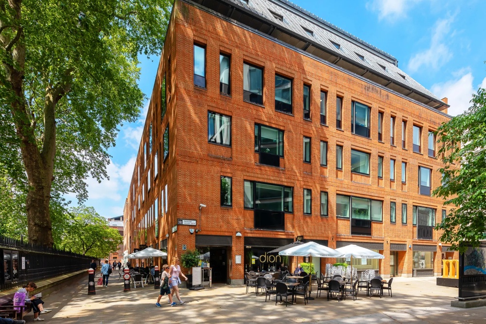 Rotes Backsteingebäude mit großen Fenstern, davor Café mit weißen Sonnenschirmen, umgeben von Bäumen.