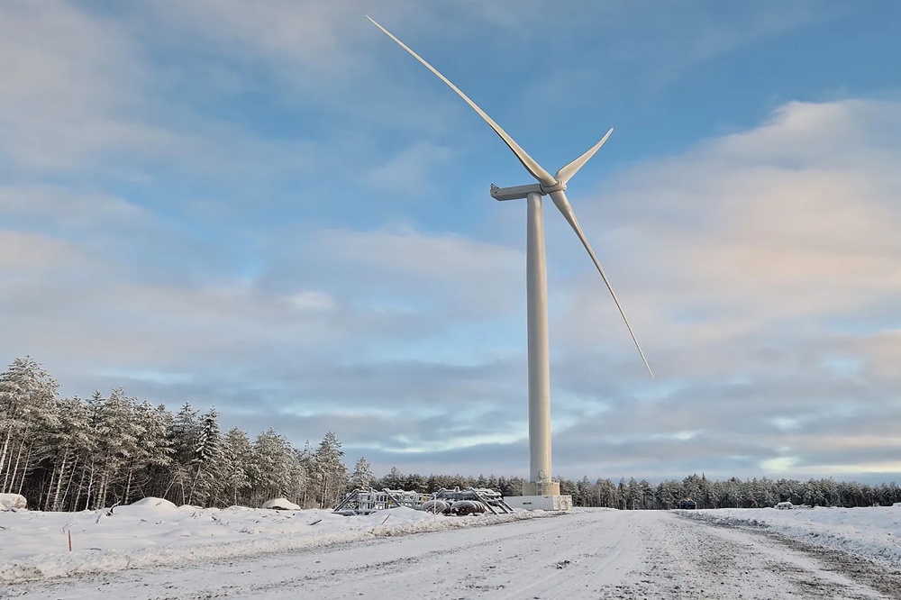 A wind turbine is standing on a snow-covered field in front of a forest and a cloudy sky.