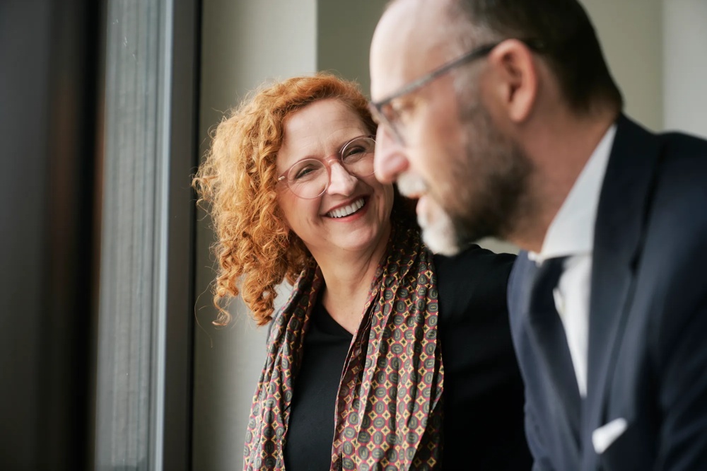 Eine lächelnde Frau mit lockigem Haar spricht mit einem Mann im Anzug vor einem Fenster.