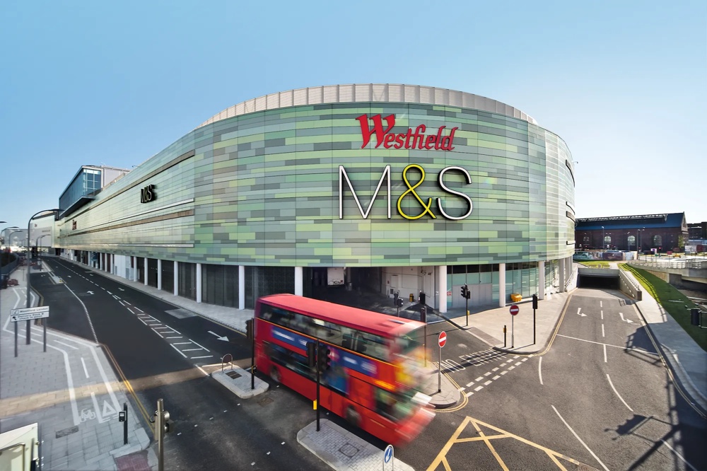 Grünes Einkaufszentrum mit M&S-Logo, roter Doppeldeckerbus auf Straße davor.