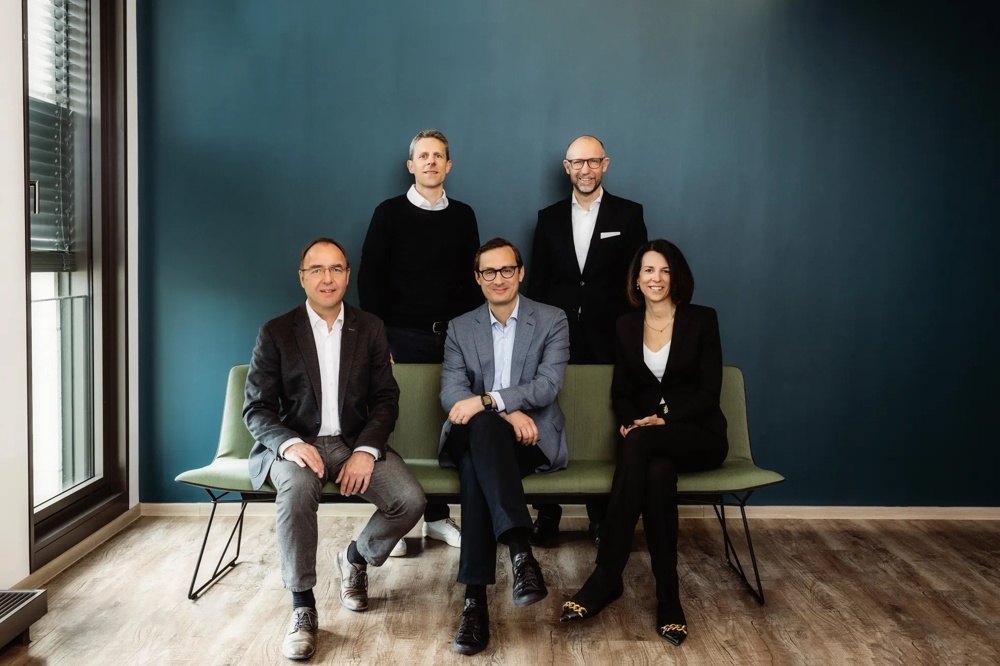 Fünf Personen in Businesskleidung posieren vor einer blauen Wand auf einer grünen Bank.