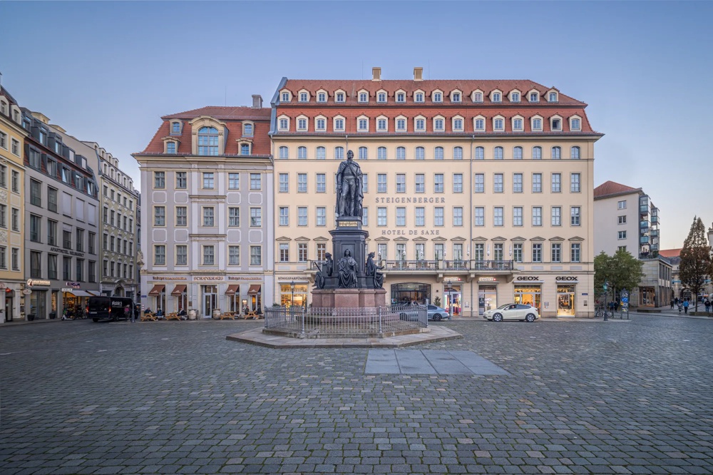 Eine Statue auf einem gepflasterten Platz vor einem großen, mehrstöckigen Gebäude mit Geschäften im Erdgeschoss.