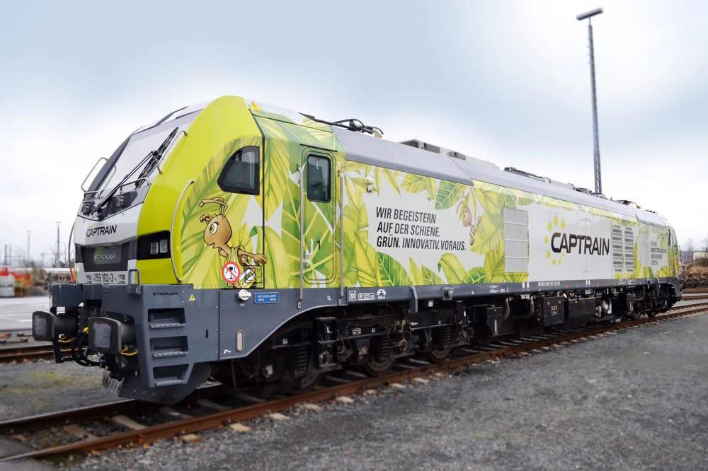 Grün-gelbe Lokomotive mit Blattdesign und Schriftzug "Wir begeistern auf der Schiene. Grün. Innovativ. Voraus." auf Gleisen.