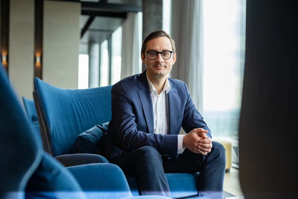 Ein Mann in Anzug und Brille sitzt auf einem blauen Sessel in einem modernen Raum.