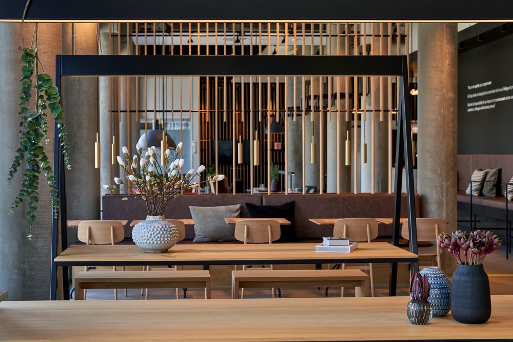 Modern room with a wooden table, vase with white flowers, hanging lamps and plants in the background.