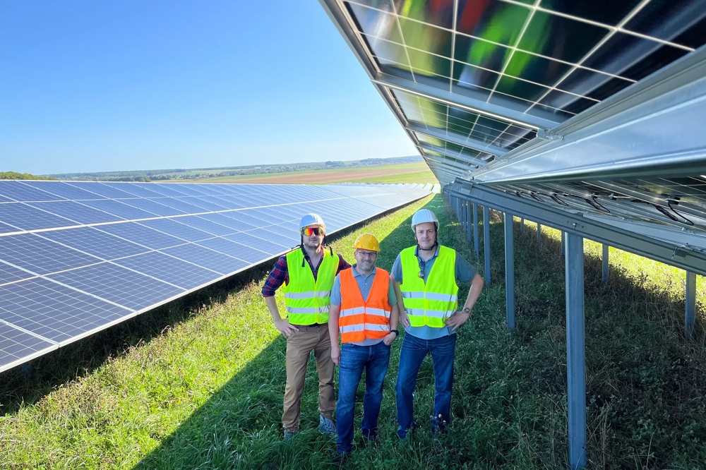 Drei Personen in Warnwesten stehen neben Solarpaneelen auf einem Feld unter klarem Himmel.