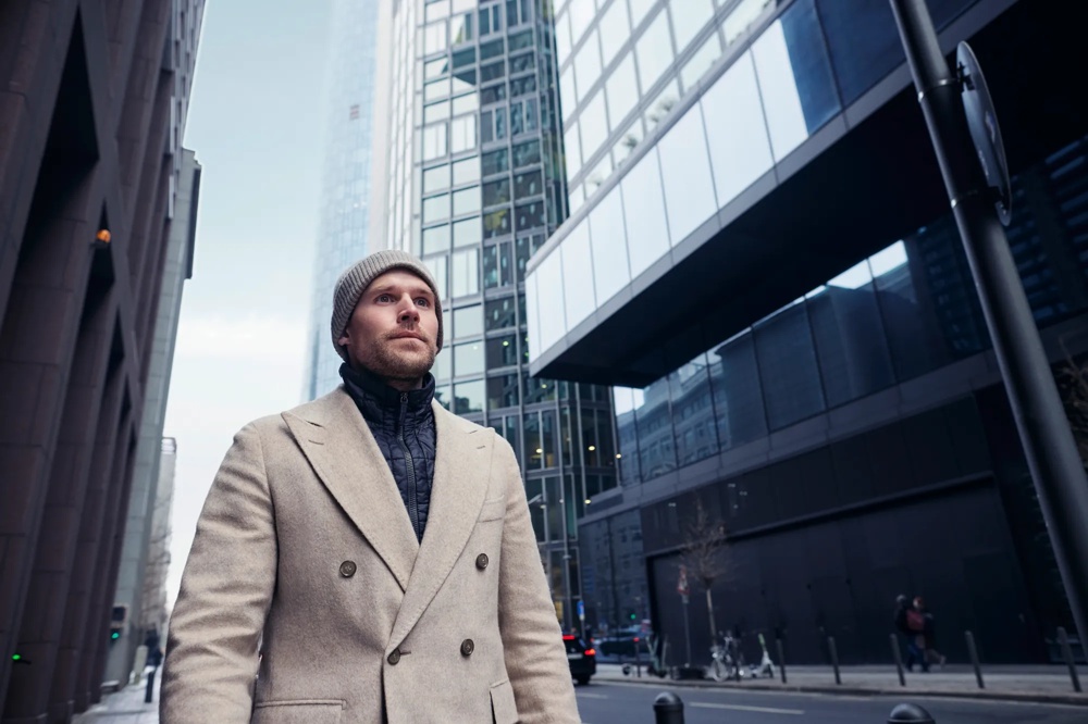 Ein Mann in beigem Mantel und Mütze steht vor modernen Hochhäusern in der Stadt.