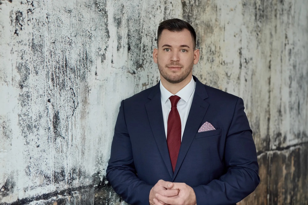 A man is wearing a black suit with a red tie and a pocket square. He is standing in front of a marbled wall. His hands are folded in front of his stomach.