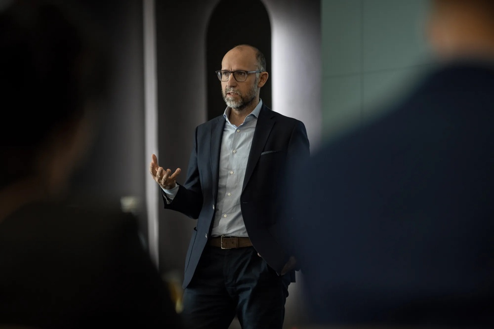 Ein Mann in Anzug spricht gestikulierend vor einem unscharfen Hintergrund.