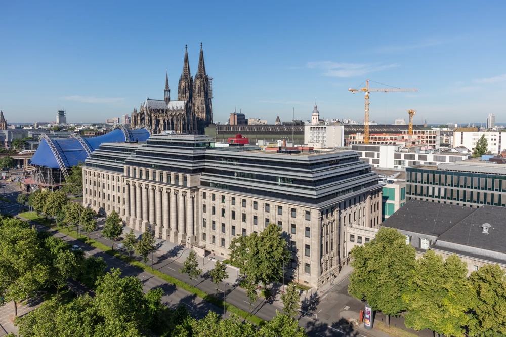 Historische Gebäude mit Kölner Dom im Hintergrund, blauer Himmel, Bäume und Baukräne.
