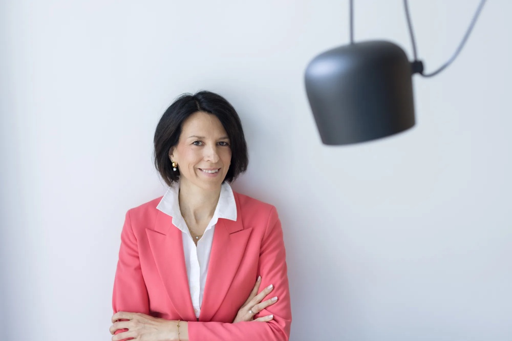 Eine Frau in einem rosa Blazer steht lächelnd vor einer weißen Wand.