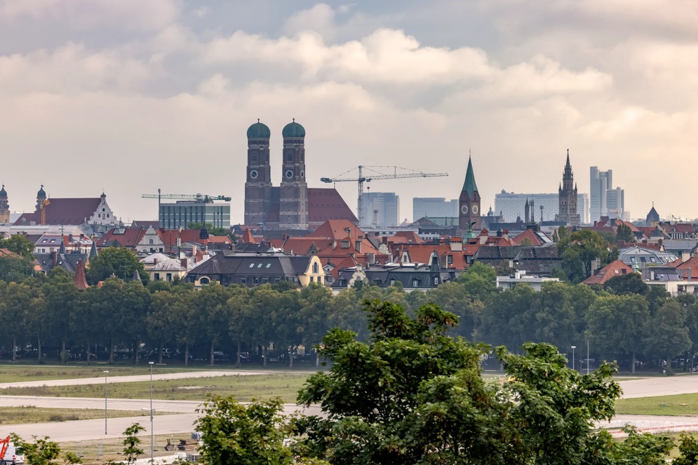 Stadtansicht mit Kirchtürmen, Baukränen und Wolkenkratzern im Hintergrund, Bäume im Vordergrund.
