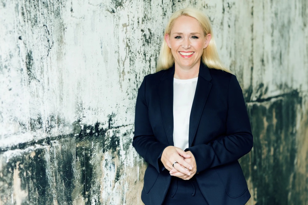 Frau in dunklem Anzug lächelt vor einer strukturierten, verwitterten Betonwand.