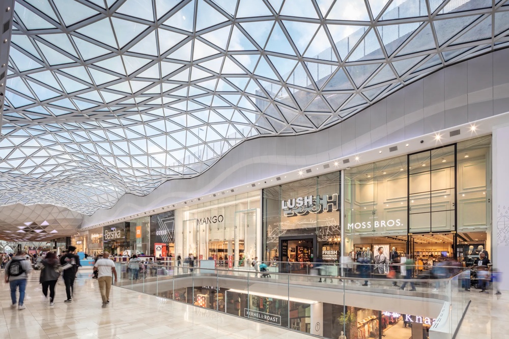 Eine moderne Einkaufspassage mit gläsernem Dach, Geschäften und Menschen, die entlang eines Geländers schlendern.