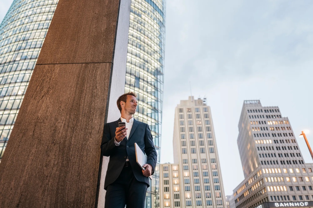 Ein Mann im Anzug steht mit Handy und Laptop vor hohen Bürogebäuden.