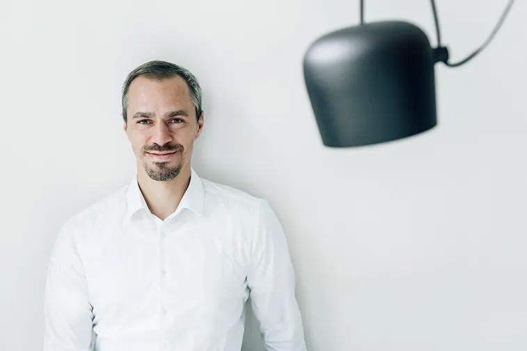 Ein Mann in weißem Hemd steht vor einer weißen Wand, neben einer schwarzen Hängelampe.