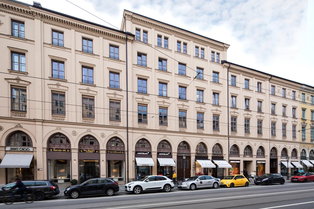 Elegantes, mehrstöckiges Gebäude mit Arkaden und Schaufenstern, davor parkende Autos und Passanten.