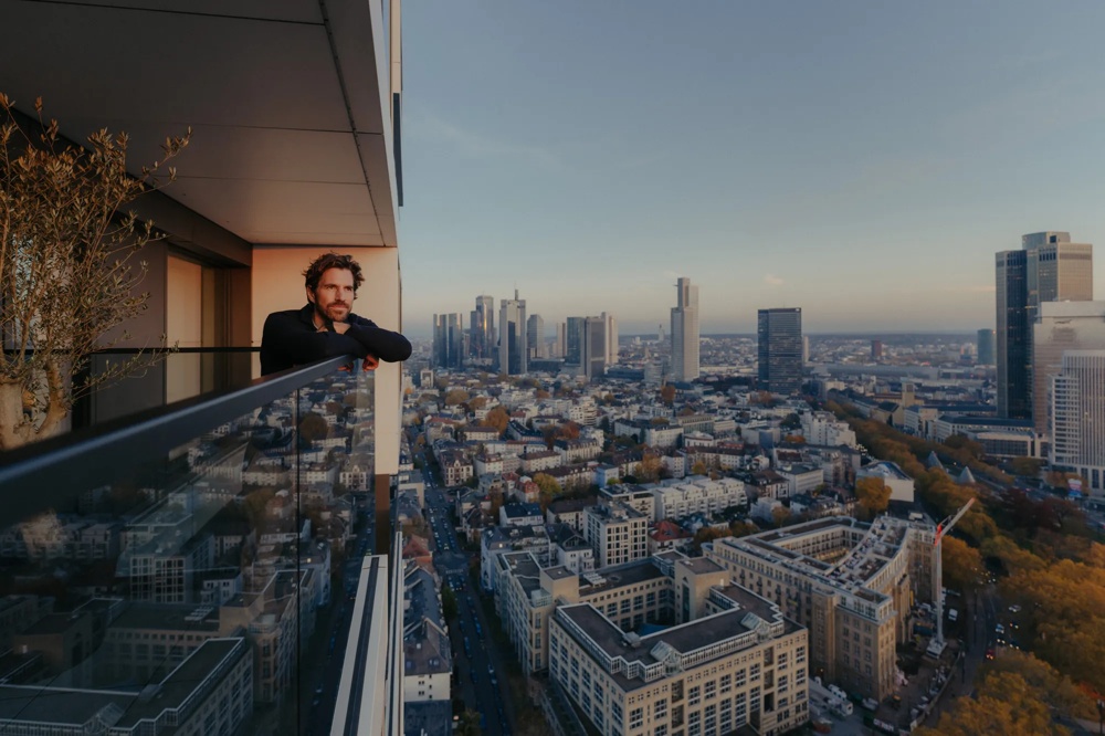 Person auf Balkon einer modernen Wohnung mit Blick über die Stadt und Getränk in der Hand.