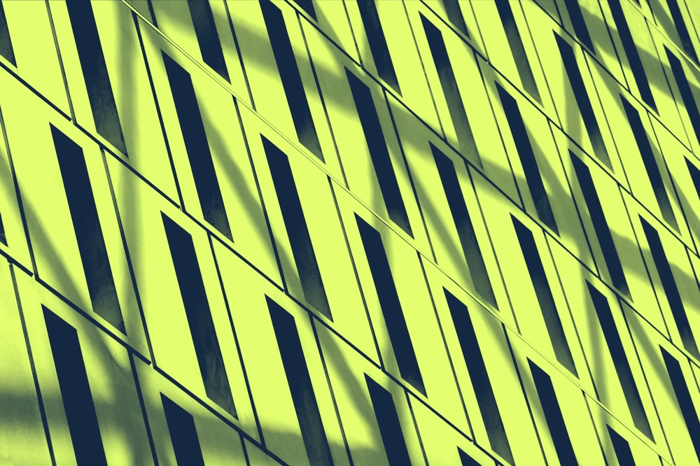 Gelbe Wand mit diagonalen, schmalen, dunklen Fenstern im geometrischen Muster, Schatten fallen schräg über die Oberfläche.