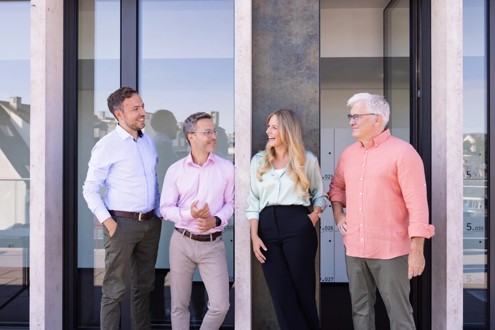 Four people smiling in front of a modern building with a glass facade.