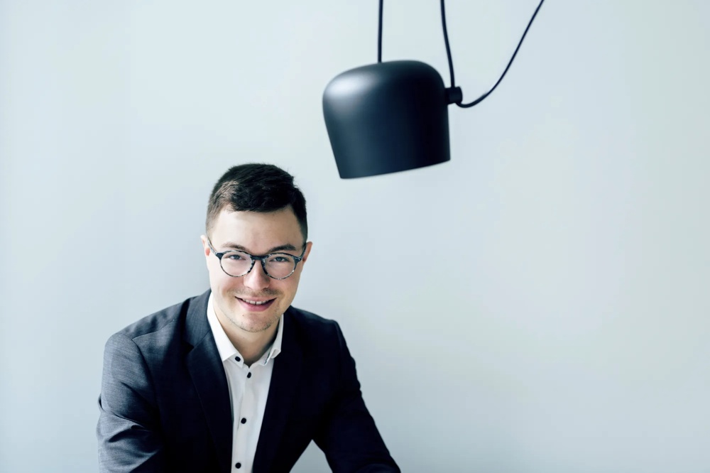 Ein lächelnder Mann mit Brille im Anzug sitzt unter einer schwarzen Lampe vor hellem Hintergrund.