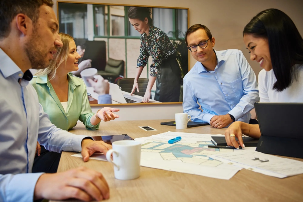 Four people discussing at a table with plans and laptops, with a large photo in the background.