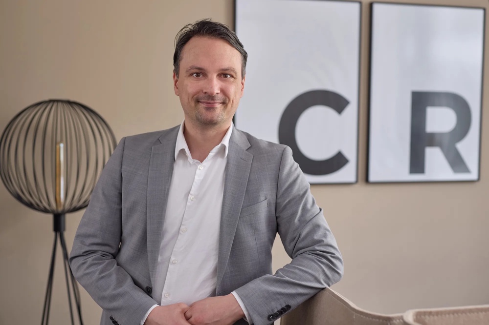 A man in a grey suit stands smiling in front of two framed letters on the wall.