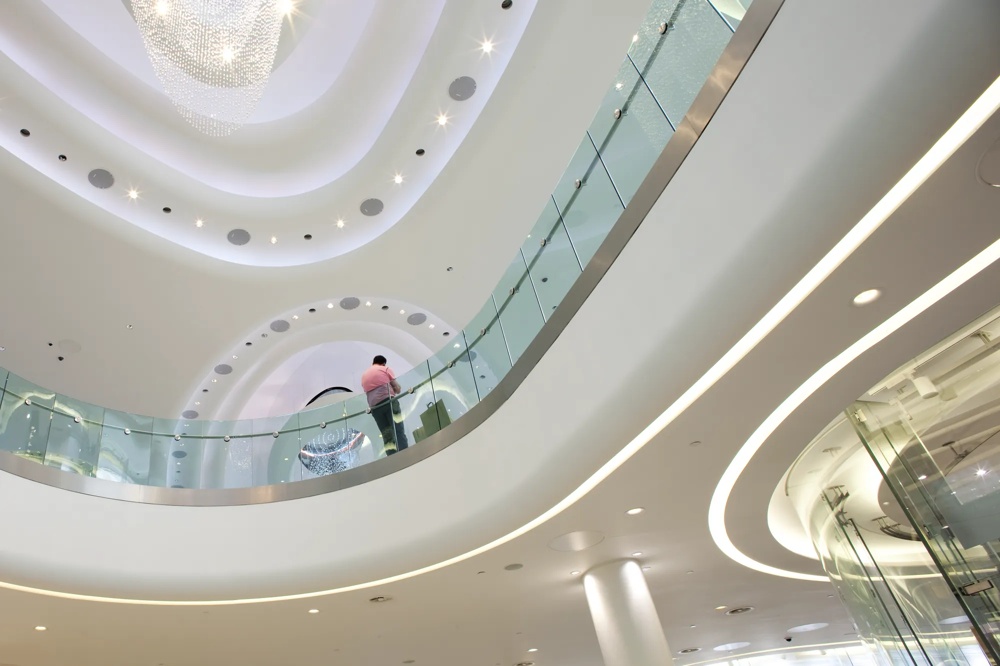 Moderne Innenarchitektur mit geschwungenen Decken, Glasgeländern und einem Kronleuchter. Eine Person in Pink steht oben.