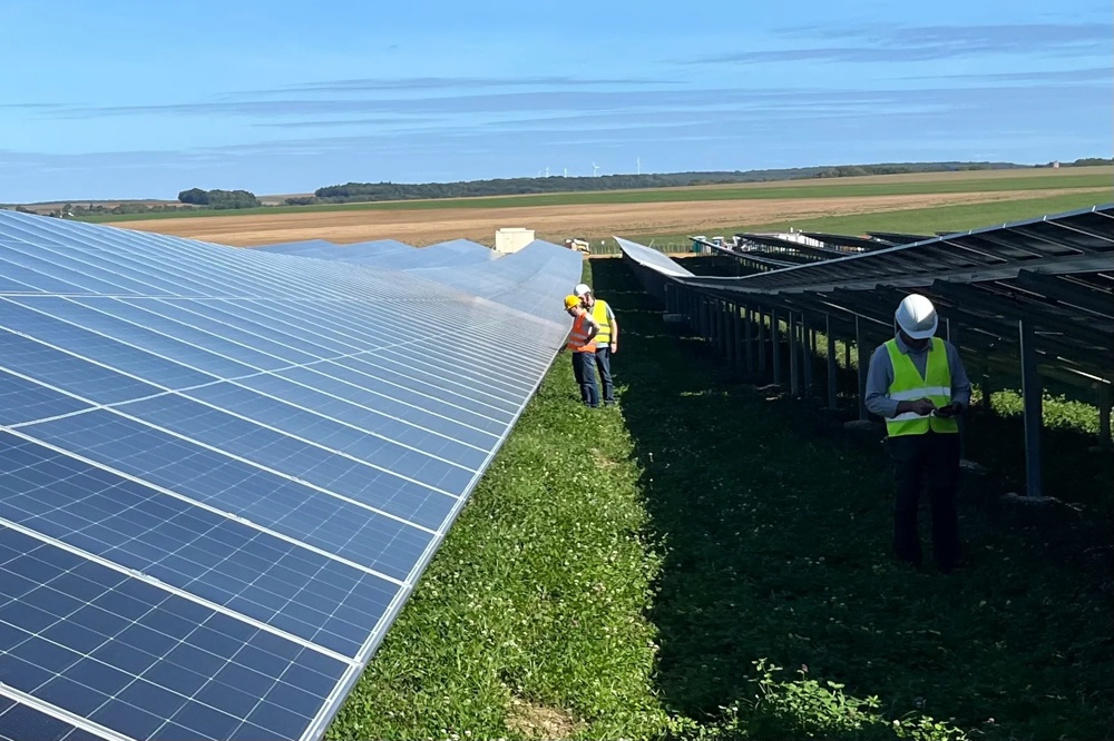Zwei Personen in Warnwesten inspizieren Solarpanels auf einem grünen Feld unter blauem Himmel.