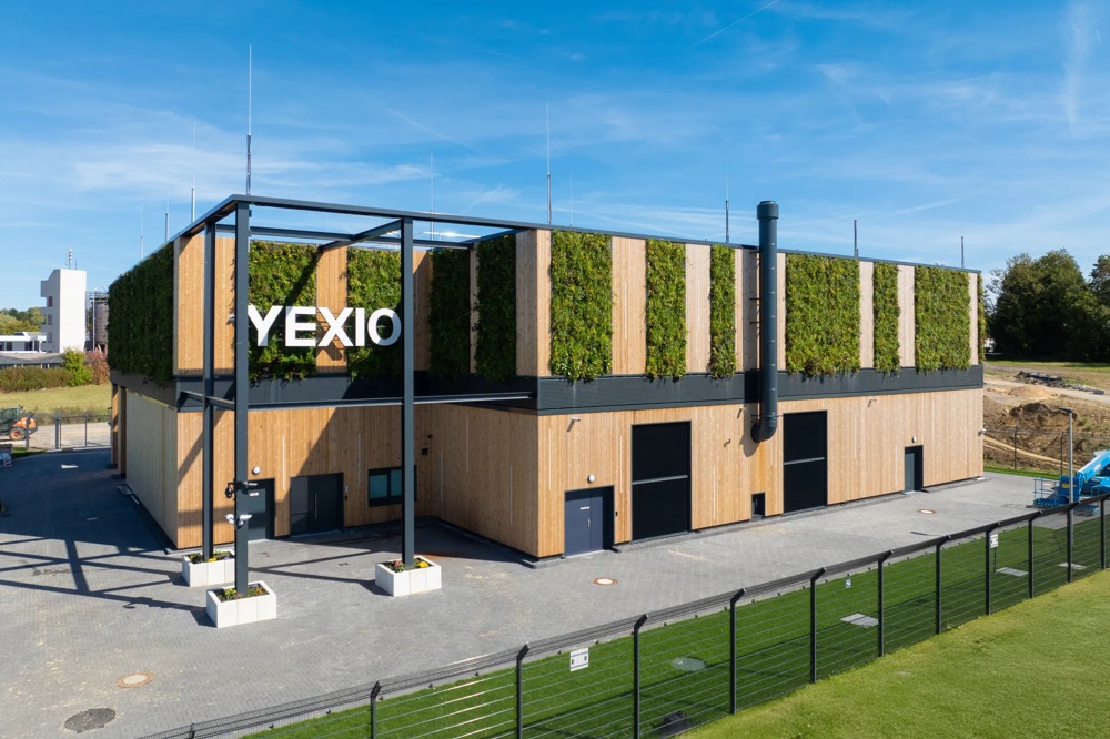Ein modernes Gebäude mit Holzfassade und begrüntem Dach neben einem Zaun und einer Grünfläche.