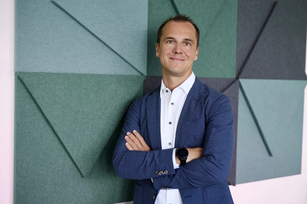 A smiling man in a blue suit standing in front of a geometric wall with green and grey triangles.
