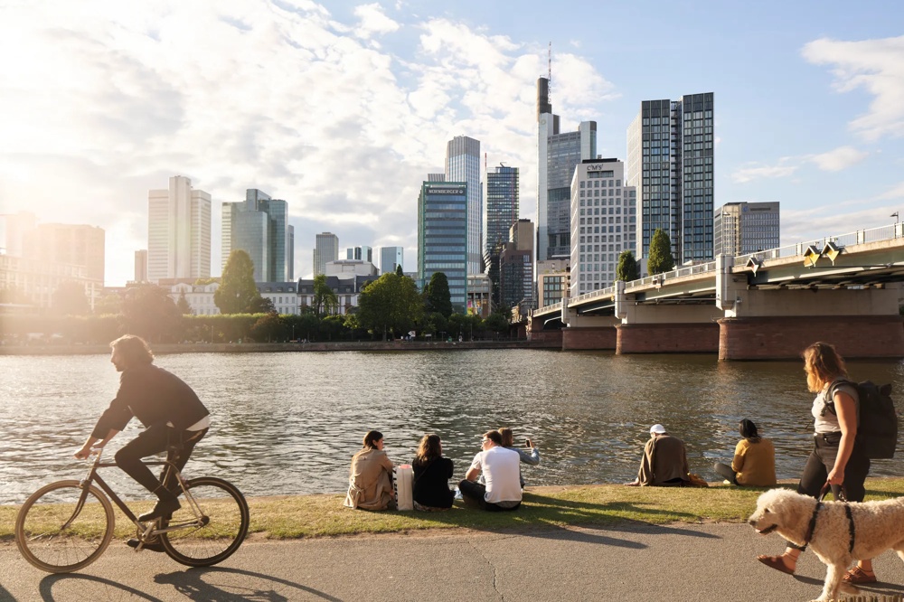 Personen sitzen am Flussufer, ein Radfahrer und eine Frau mit Hund im Vordergrund, Hochhäuser im Hintergrund.