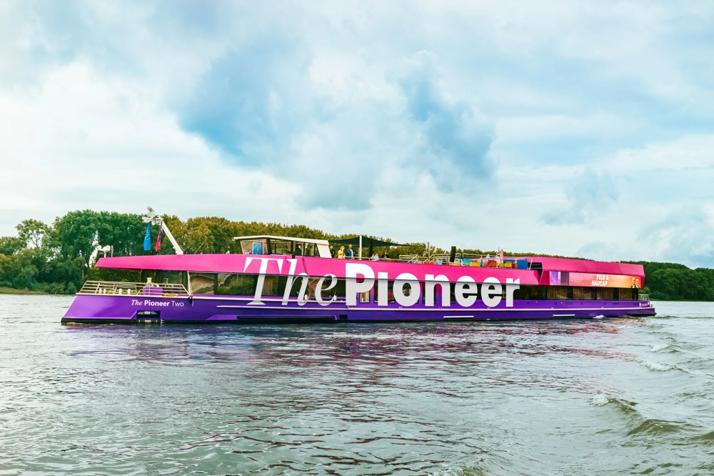 Ein pink-lila Boot mit der Aufschrift "The Pioneer" fährt auf einem Fluss, Bäume im Hintergrund.
