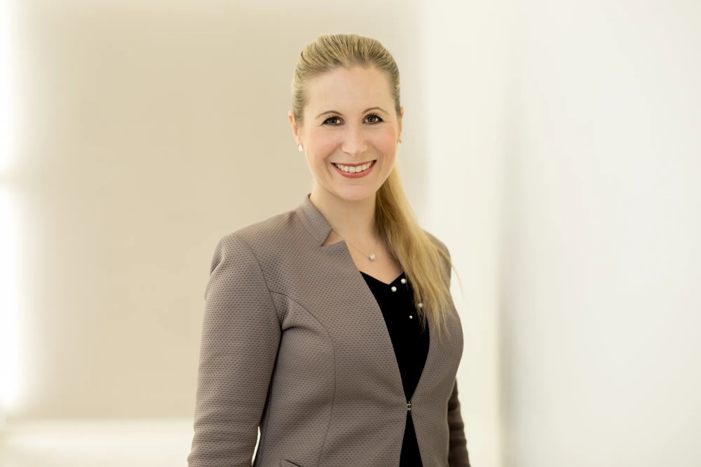 A smiling woman with a blonde ponytail in a grey blazer against a light background.