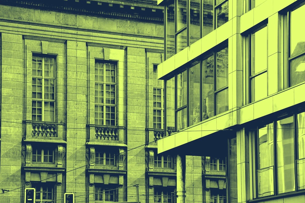 Moderne Glasfassade trifft auf historische Steinarchitektur in gelb-grünem Farbton.