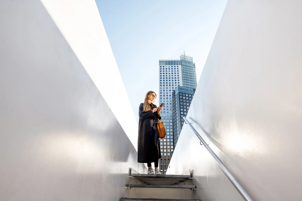 Woman with mobile phone on stairs, high-rise building in the background, blue sky.