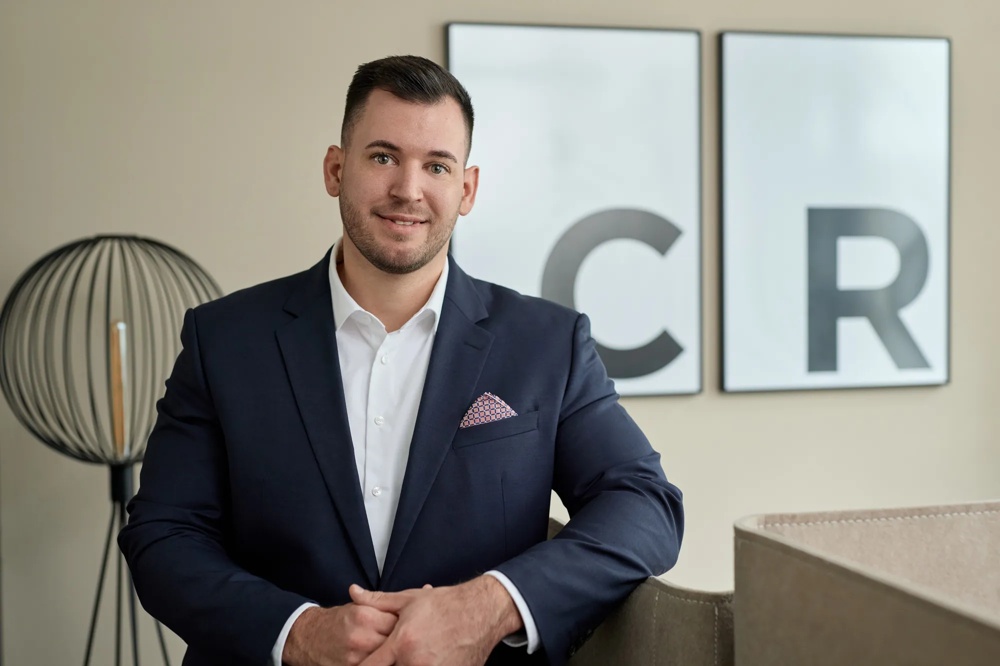 Ein Mann trägt einen schwarzen Anzug ohne Krawatte und ein Einstecktuch. Er steht mit einem Arm auf einer Abgrenzung gelehnt vor den gerahmten Initialen der Commerz Real und einem Lampenschirm.